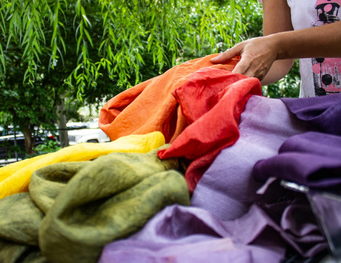 The hands and body of a participant is on the right of frame, facing left, and holding a pile of materials. The materials are the dyed silk to be cut into flowers for the puppet. The materials are red, orange, green, yellow, and purple. Due to COVID, this event was held outside, so there are green trees in the background. This is is NYC.