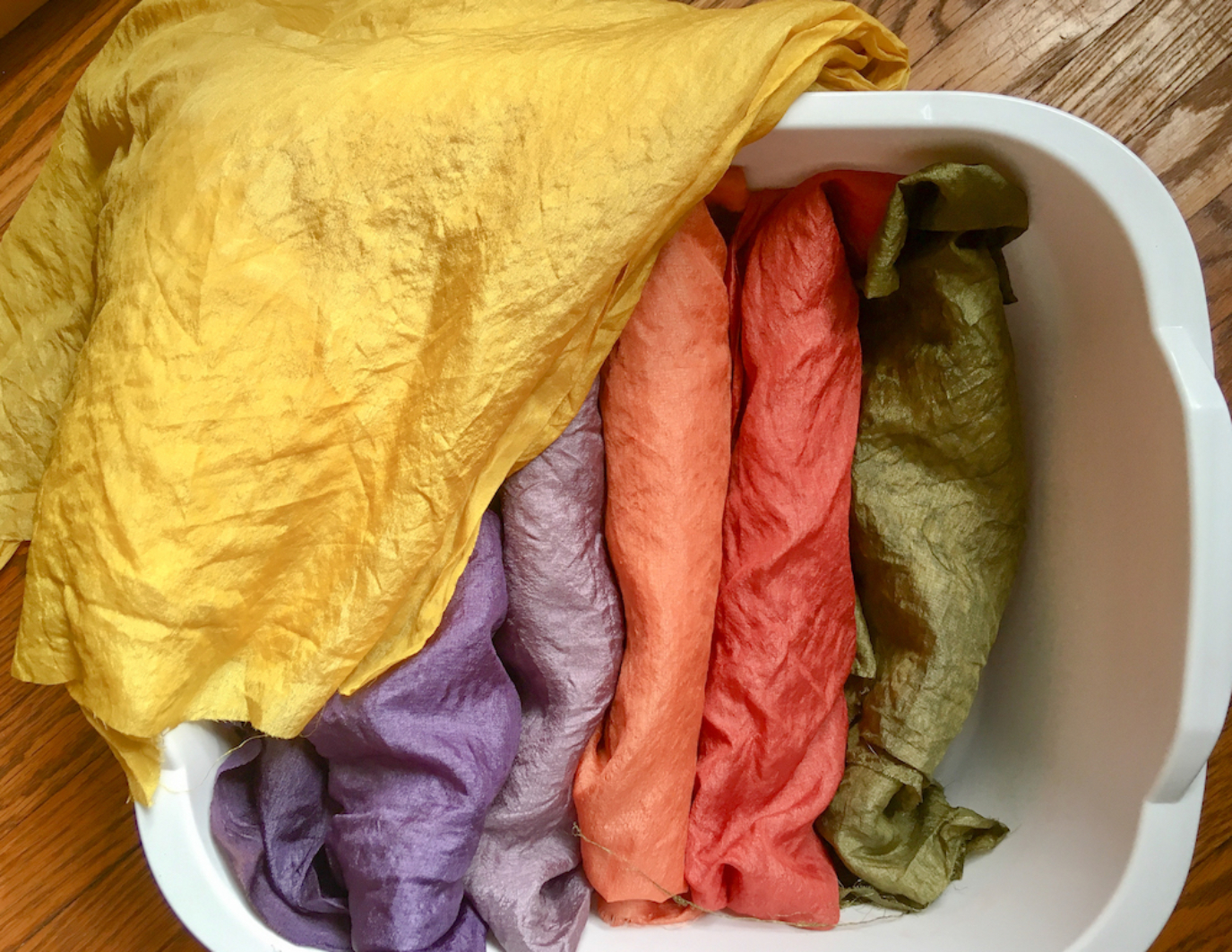 A white plastic tub filled with hand dyed fabrics rolled up to be used for the puppet. Six silks rolled and placed side by side, left-right: yellow draping over a corner of the bucket, with purple, lavender, orange, red, and green.