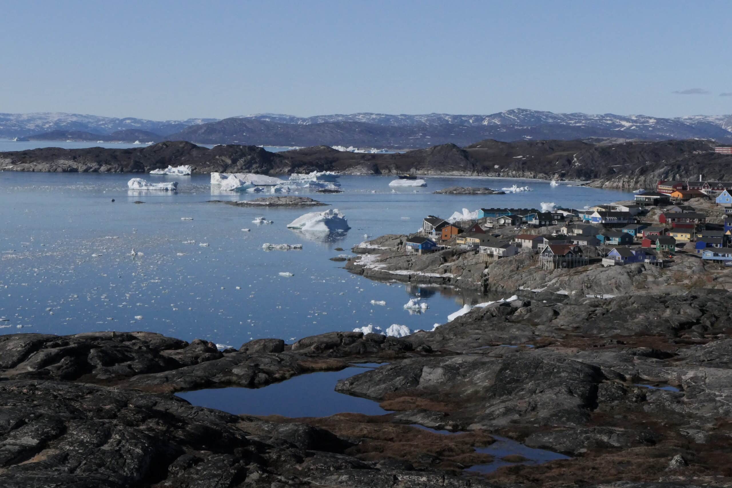 Rocky land dotted with colorful houses rings water that has icebergs floating in it. 
