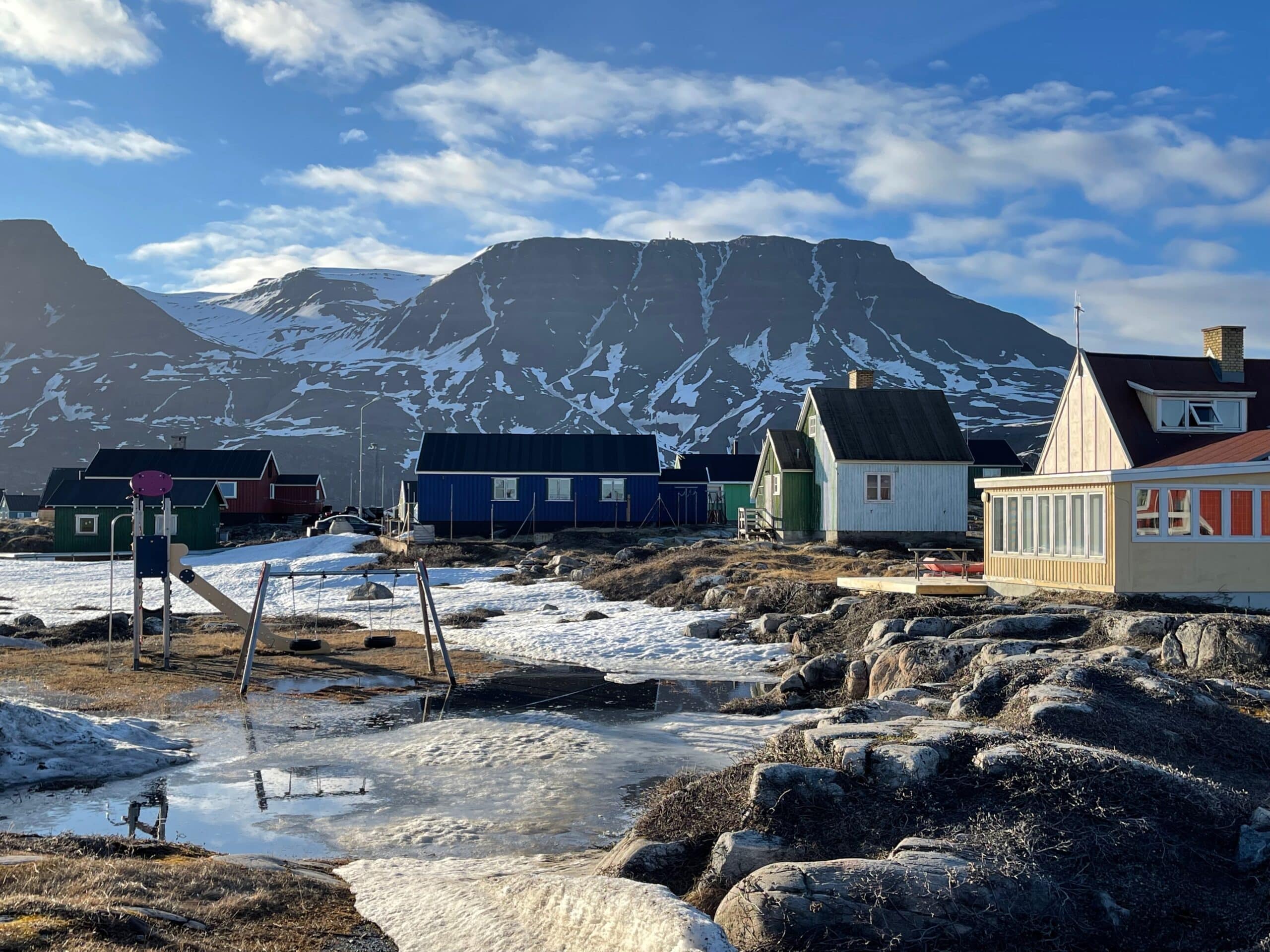 A small cirlce of colorful houses encircle a small playstructure on rocky ground and melting snow and ice. Snowy mountains rise behind them.