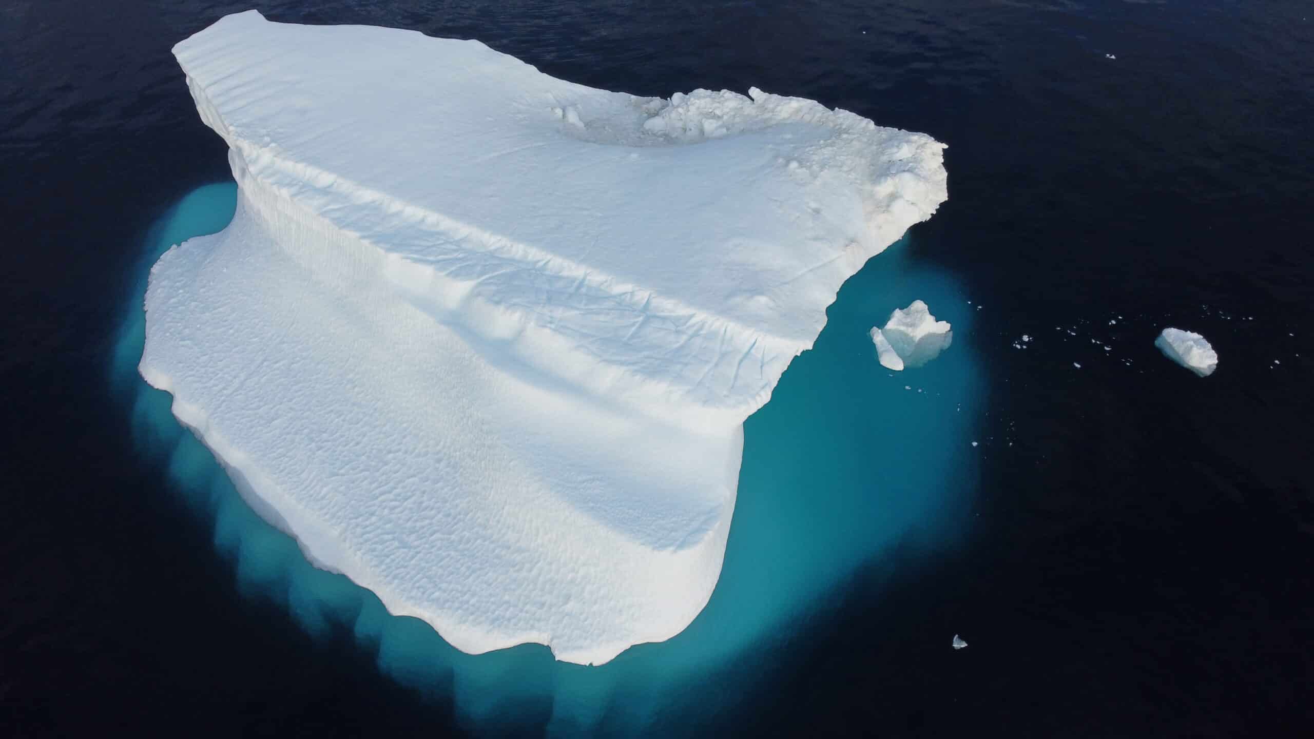 A white iceberg surrounded by dark water. The ice below the surface appears turquoise. 