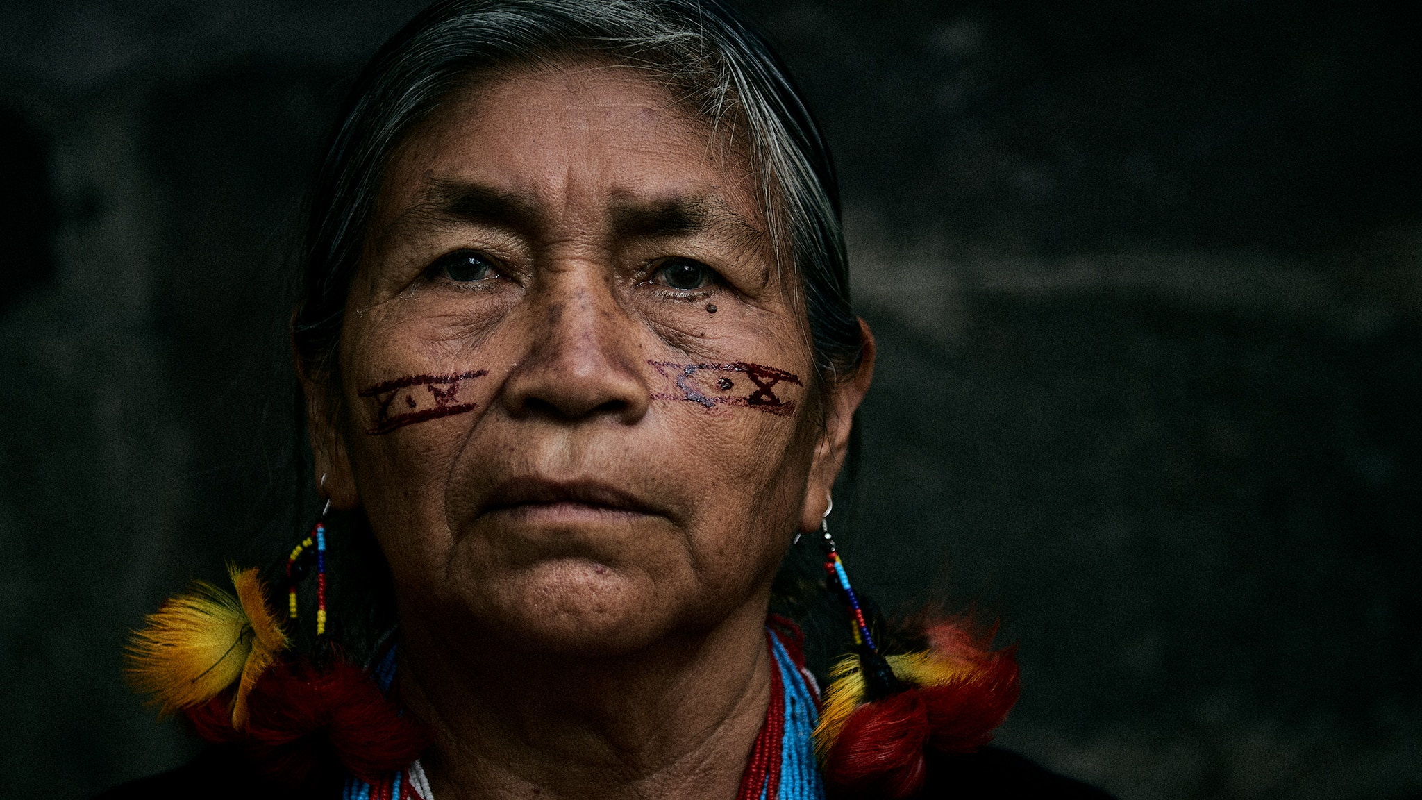 A close-up image of Catalina Chumpi. She is an older Indigenous woman with painted markings on her face. She wears beaded necklaces and bead and feather earrings. 