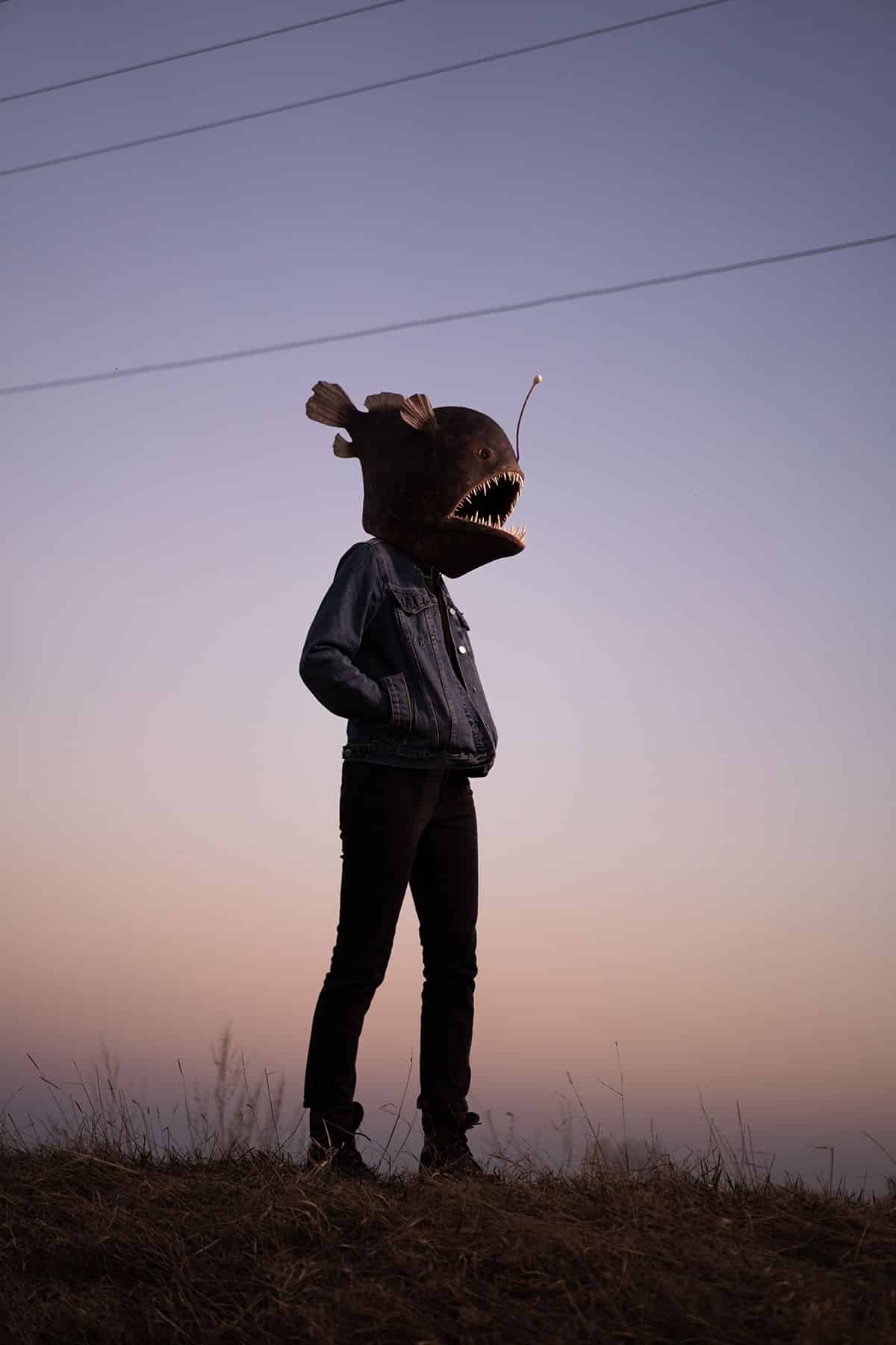 A person wearing a denim jacket at dusk with an angler fish head over their head
