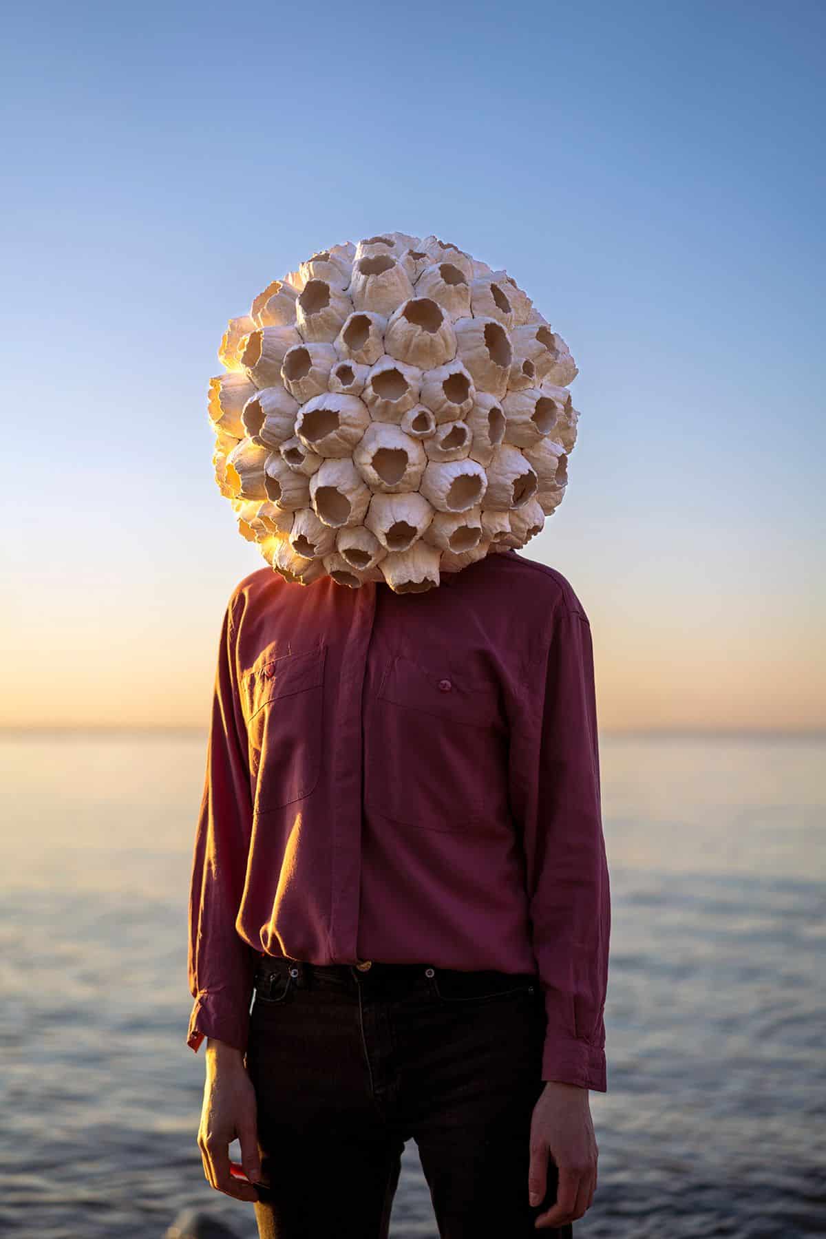 A person wearing a burgundy shirt and black jeans standing on the edge of water wearing a sphere of barnacles over their head