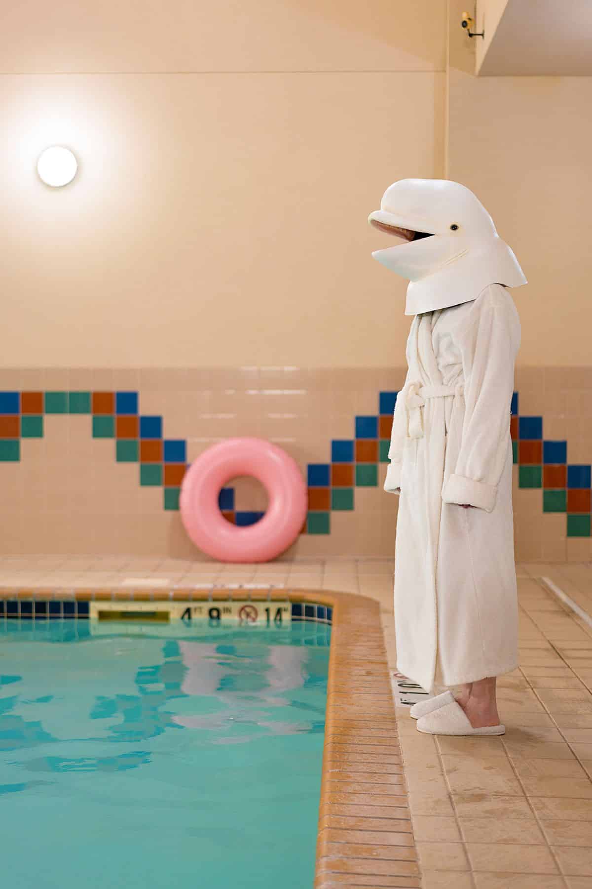 A person standing on the edge of a hotel pool, wearing a white robe and the head of a beluga whale over their head