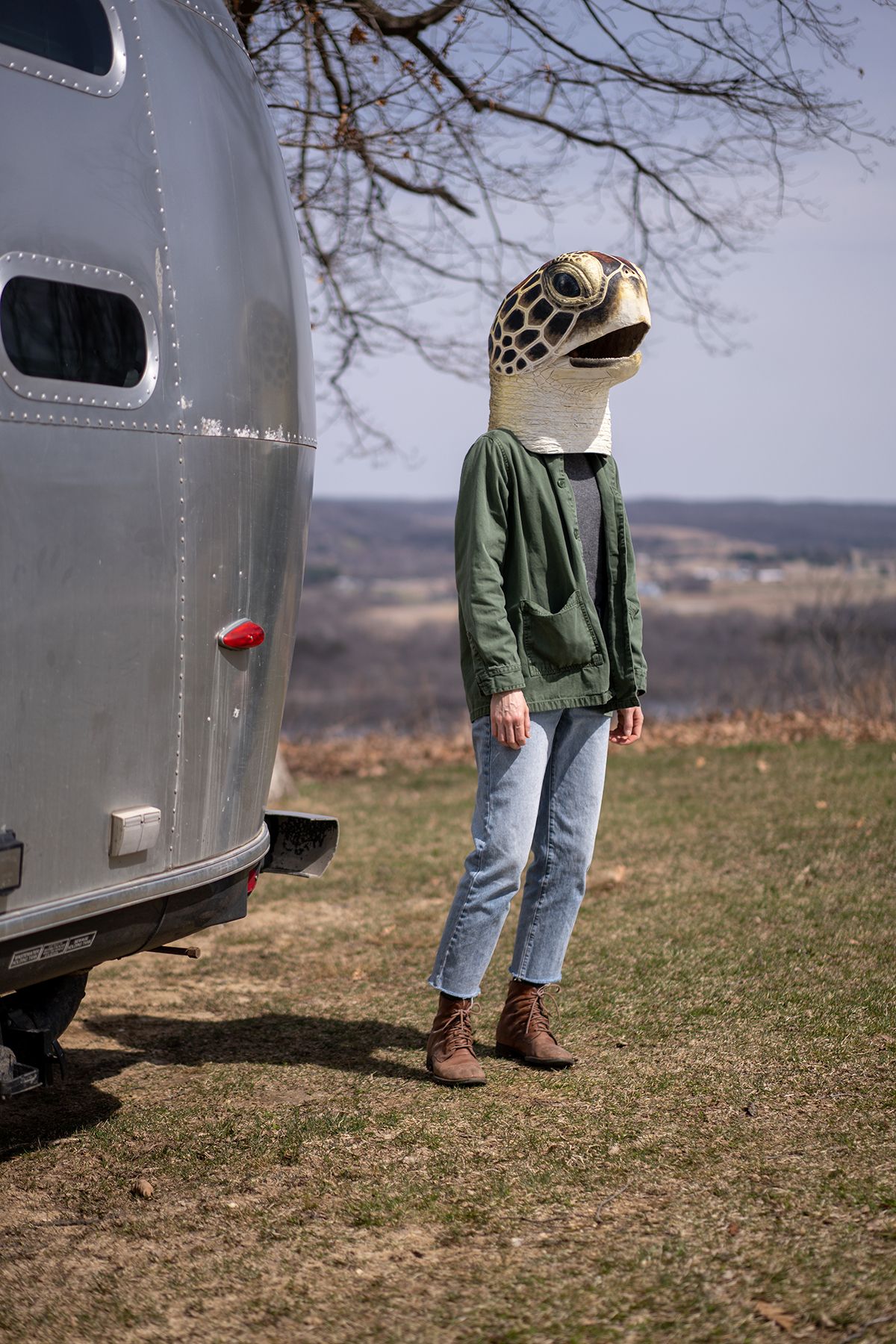 A photo of a person in a green jacket and blue jeans standing next to a silver trailer while wearing a sea turtle head over their head