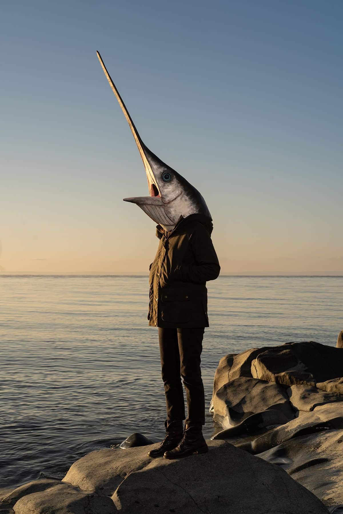 A photo of a person standing on rocks on a seaside wearing a swordfish head on their head