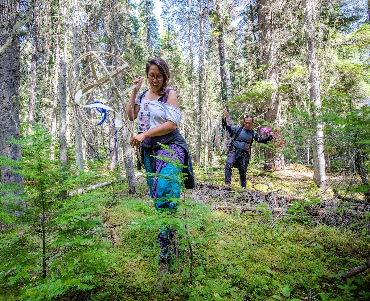 The artist Marina Tsaplina and her partner walk in an overgrown forest in Montana's Yaak Valley. Marina leads, and is highstepping through thick undergrowth and dead fallen trees. The understory is green and verdant, and blue sky appears in the background through thick trees. The artist is smiling and both of them are holding components of the Dream Puppet, which are a structure of bamboo and pink/purple/yellow flowers.