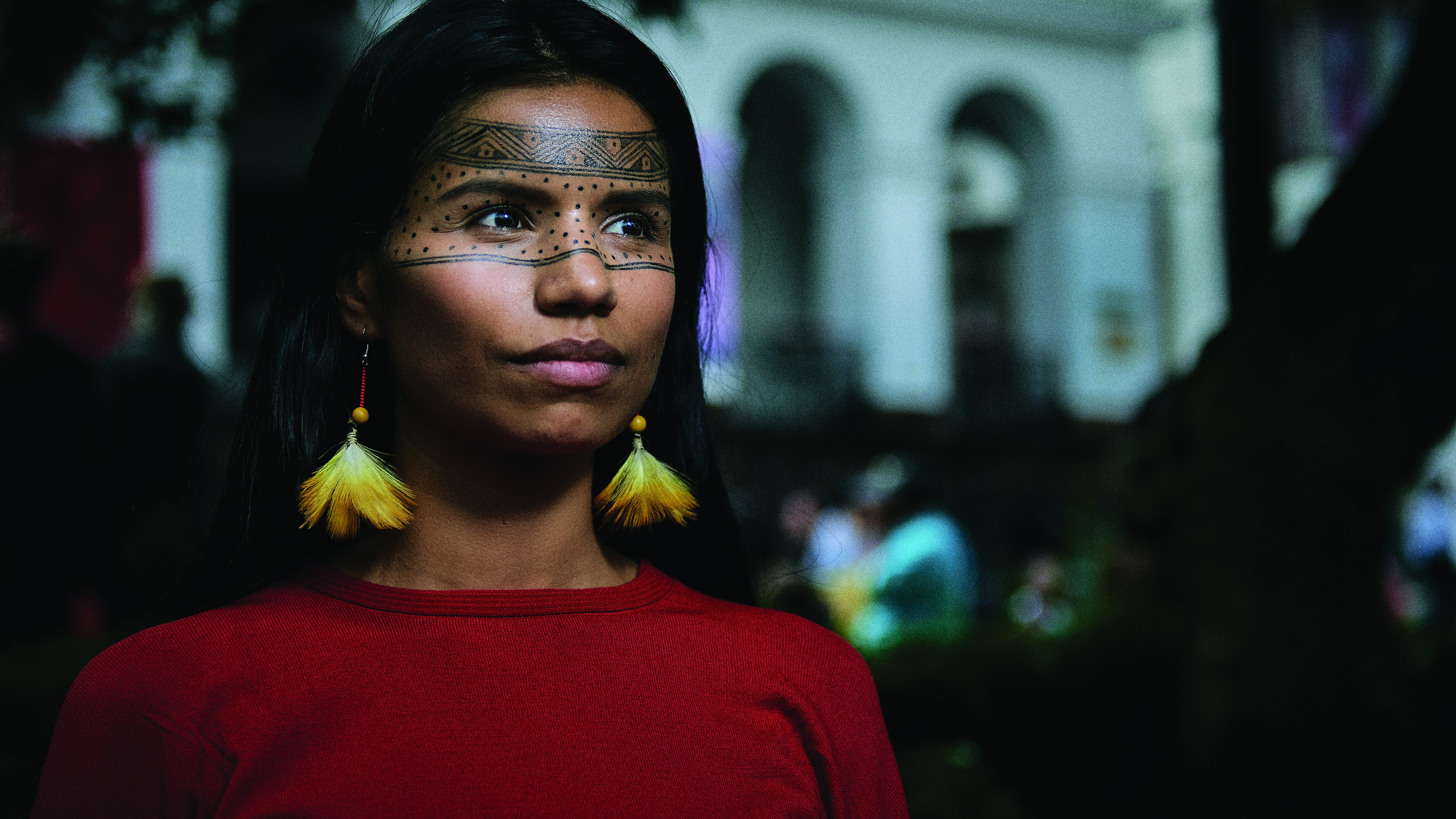 A close-up image of Nina Gualinga. She is an Indigenous woman with painted markings on her face. She wears bead and feather earrings and her shirt is red. 