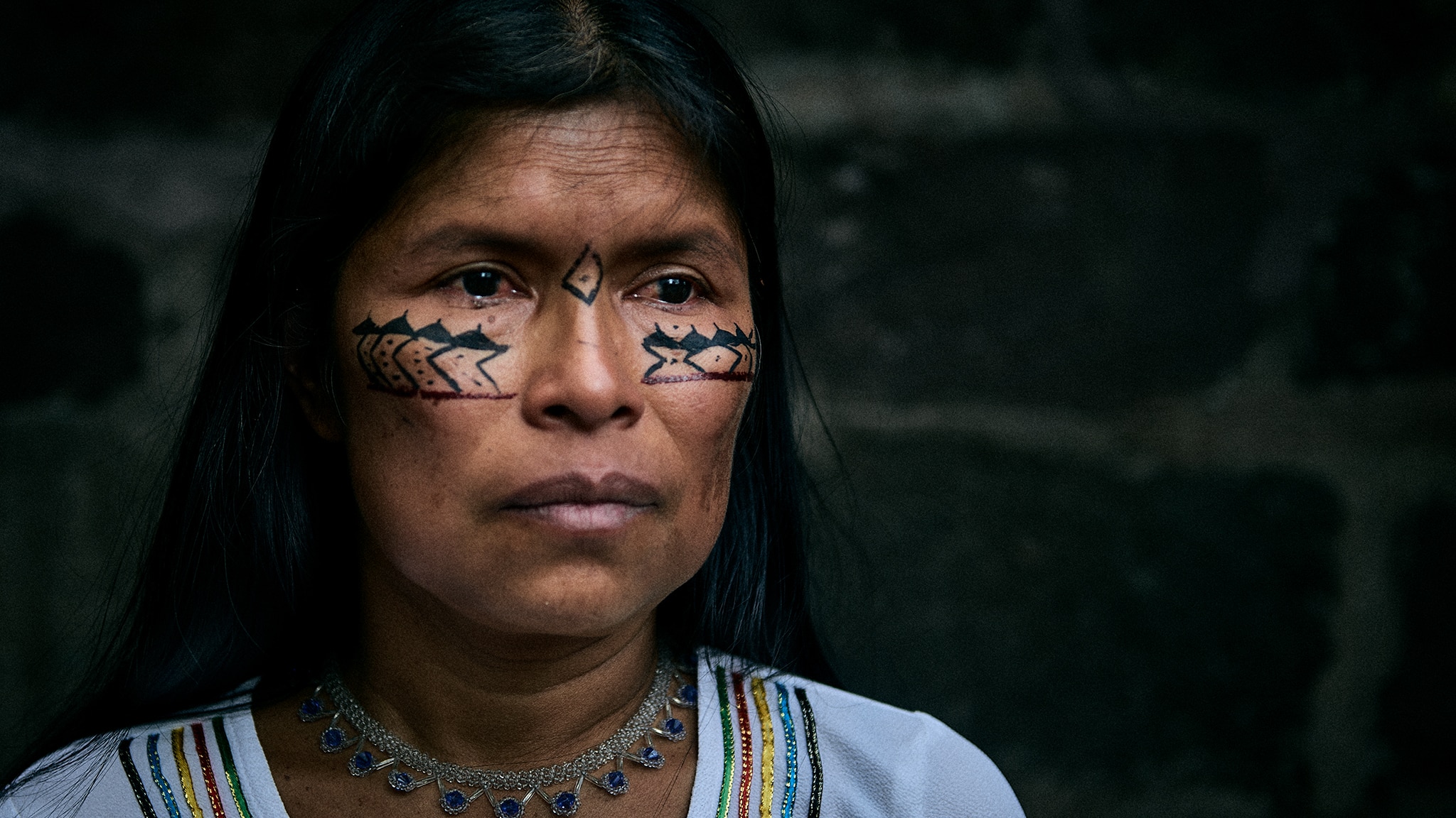 A close-up image of Noemi Gualinga. She is an Indigenous woman with painted markings on her face. She wears a necklace made of metal and blue stones. Her shirt is white with green, red, yellow, blue, and black stripes around the neckline. 