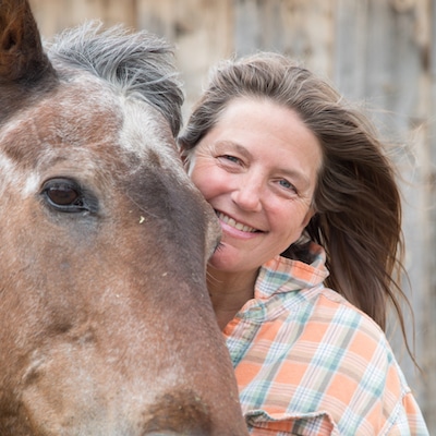 Pam Houston is on the right side and her horse on the left. The horse is brown and Pam is wearing an orange, white, and blue/gray checkered plaid shirt. Her hair is brown, like the horse, and she is smiling.