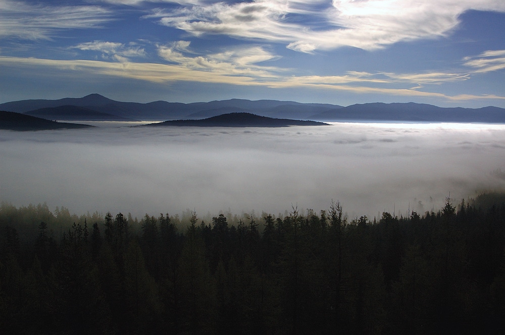 A fog sits over much of a wilderness forest, with dark mountains in the background and a blue sky with wispy clouds. Mountains poke through the fog like islands.