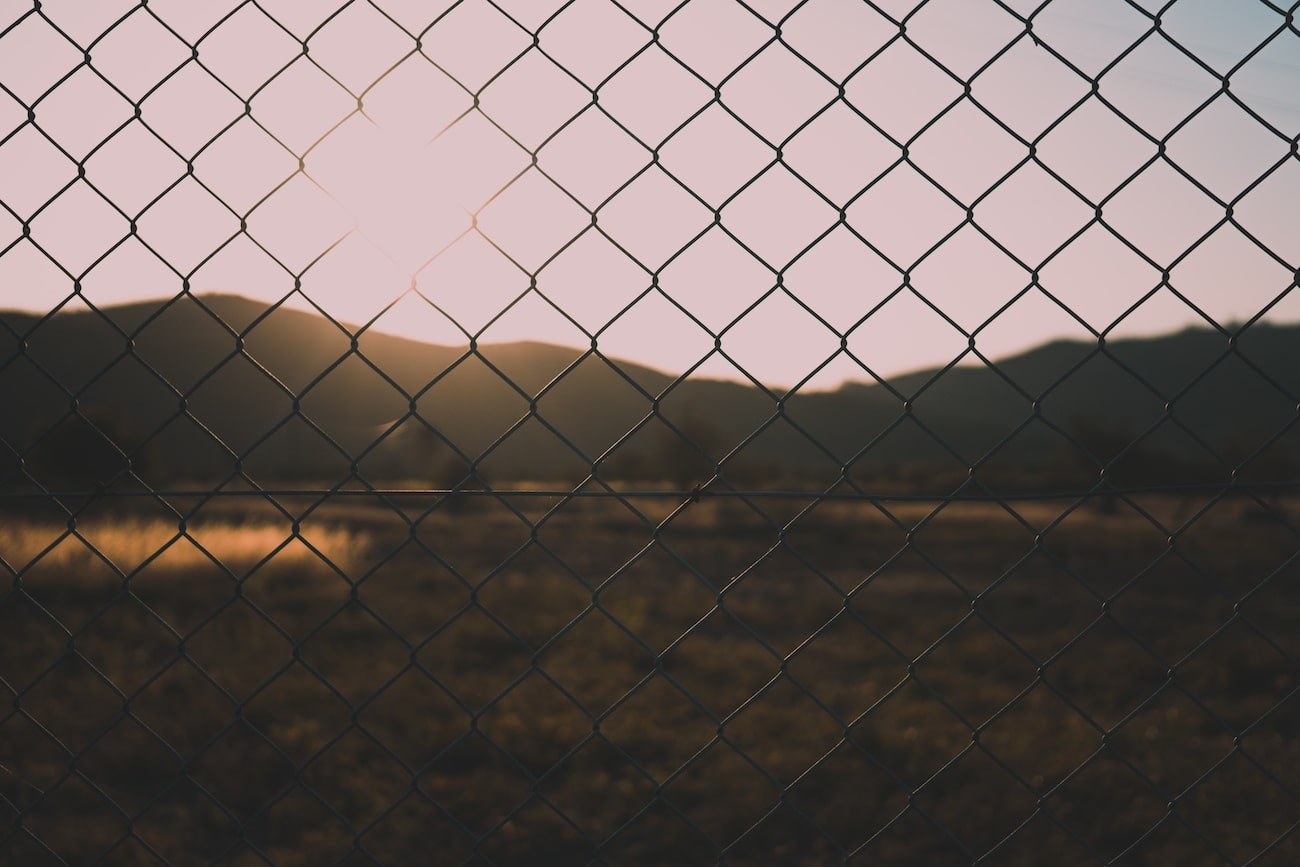 A chain link fence transposes in front of a natural space behind. A mountainous landscape with the sun shining on the left third of the sky. The ground is undeveloped and brown-green.