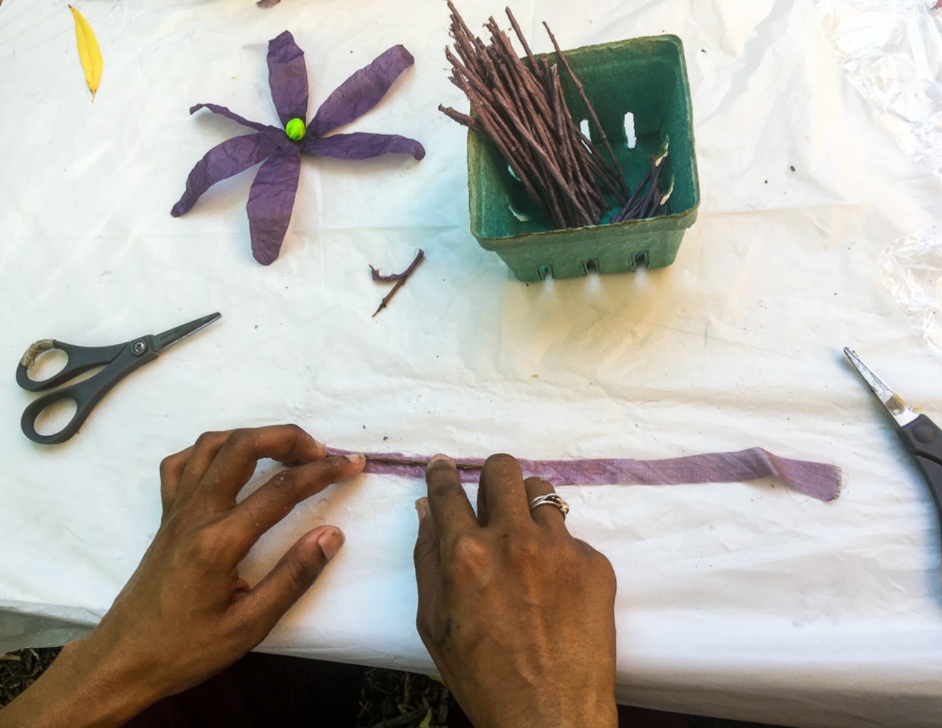 A pair of hands at the bottom of the frame is building purple flowers on a white table. There is a pair of scissors, a little box of sticks, and one completed flower (has a green button in middle of six petals.) This is the NYC event.