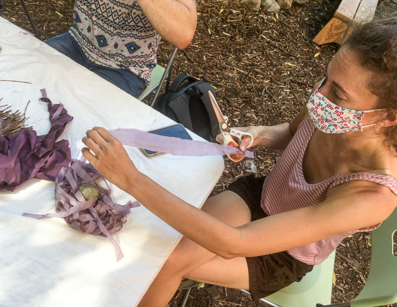 One artist/volunteer is cutting strips of lavender colored materials. She is masked and wears a red striped shirt.