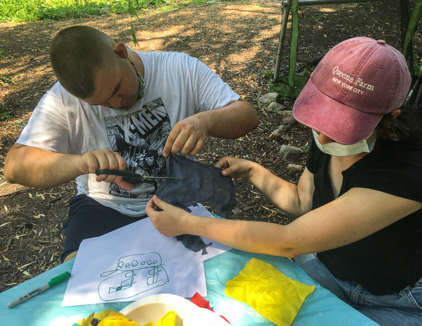 Two volunteers are working together to create a part of the Puppet. One person is holding a piece of black fabric, (she wears a red hat), while the other person is cutting into the fiber. They are both sitting at a white table with a sketch of the puppet and other yellow squares of material.