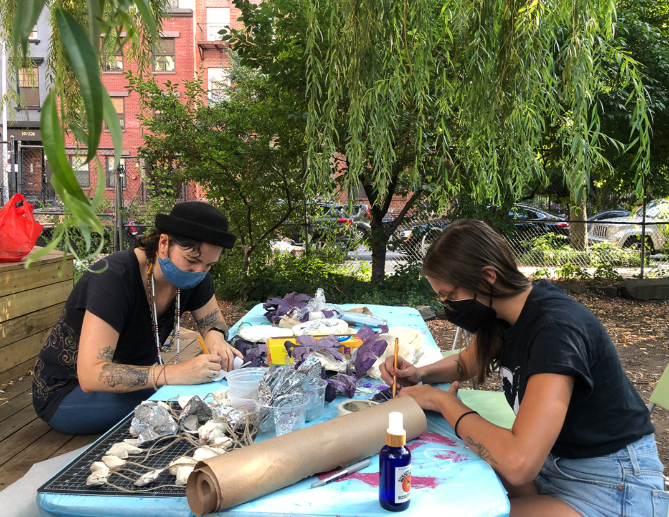Two artists sit face to face on a blue table, and they both have pencils in their hands and are writing or drawing something. The table is filled with materials for the puppet. In the background, there are trees.