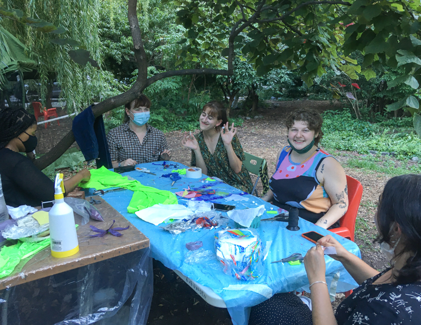 Five volunteers sit around a blue table with paints and materials, smiling and posing for the camera. Some are masked. There are trees and nature surround them. This is the NYC event. Colors on the table include green, white, red, and black