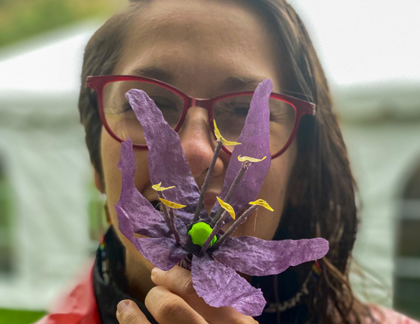 The artist, Marina, holds up in front of her face, the first completed Camas flower, a collaborative effort of hand dyeing and transport. There are six purple petals and a green knob in the middle, with several stamen with yellow flowers on them. Her face is in the background and she is smiling.