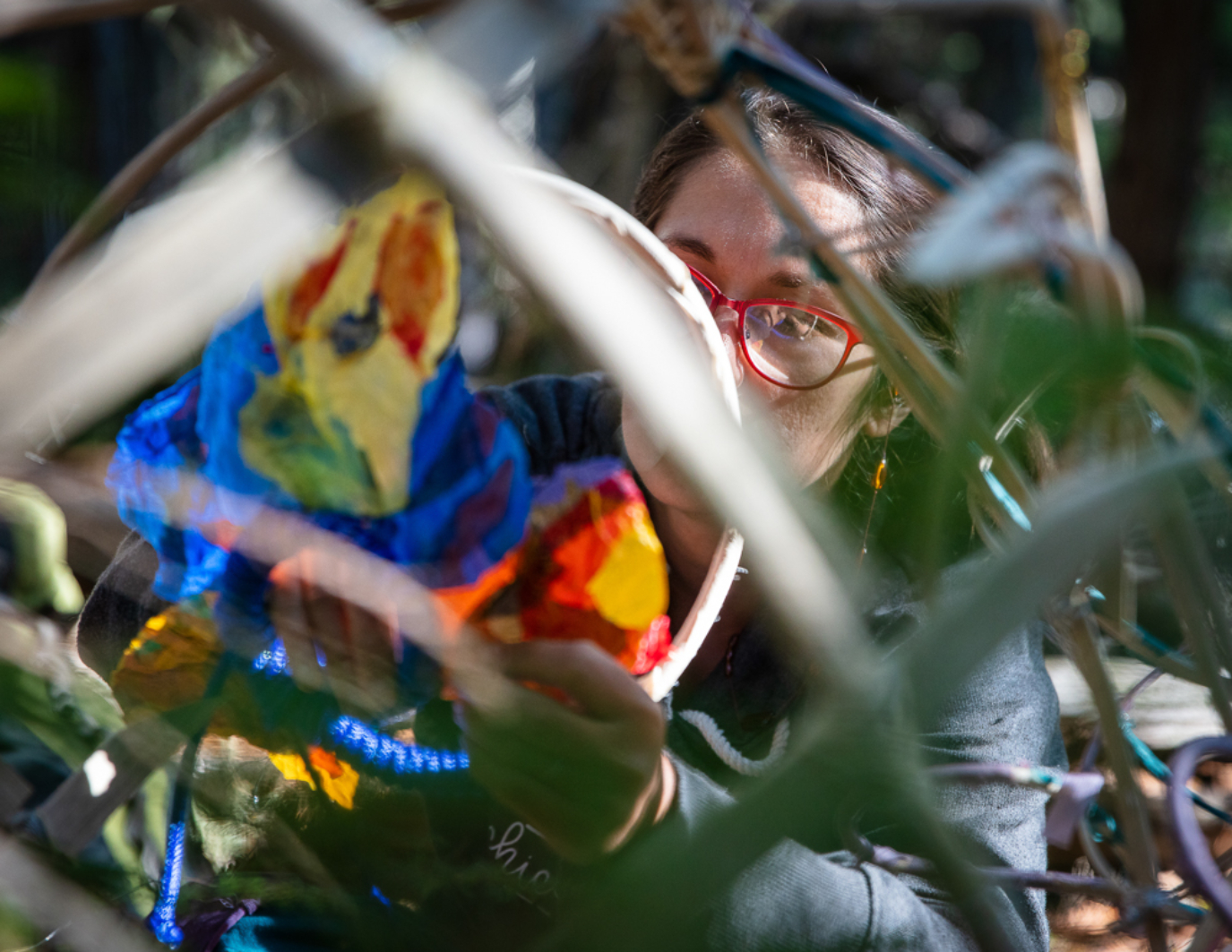 The artist face is behind a tangle of materials as she installs the puppet. She is wearing red glasses and her hands are affixing flowers to the bamboo structure.