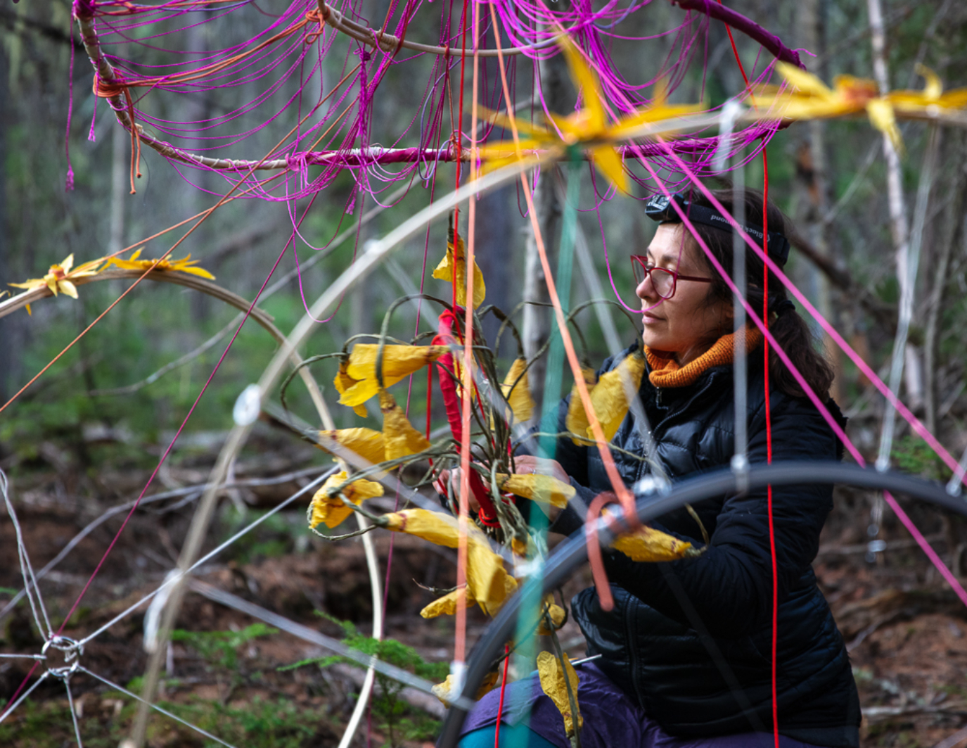 The artist sits to the right, facing left, with a smile, making last fixes to the Dream Puppet. There are bamboo arcs and curves everywhere, with yellow flowers, pink and and purple string. In the blurred background, the forest of Montana's Yaak Valley looms.