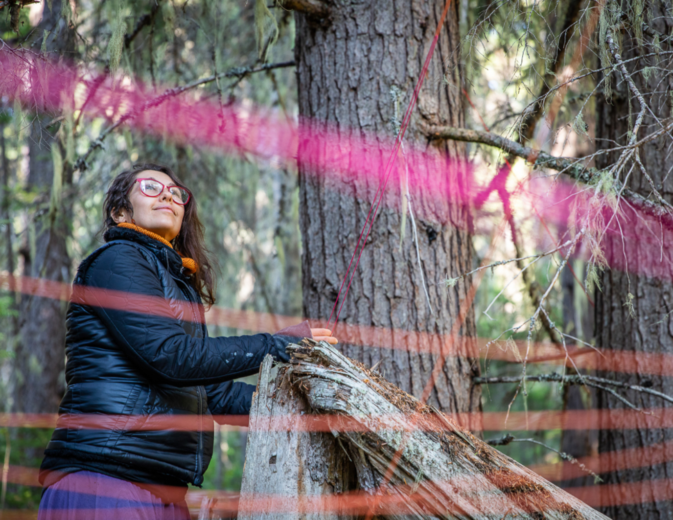 The artist looks up with a smile at the Dream Puppet, now installed in the forest. There are blurred pink and red lines that hold the puppet in place.