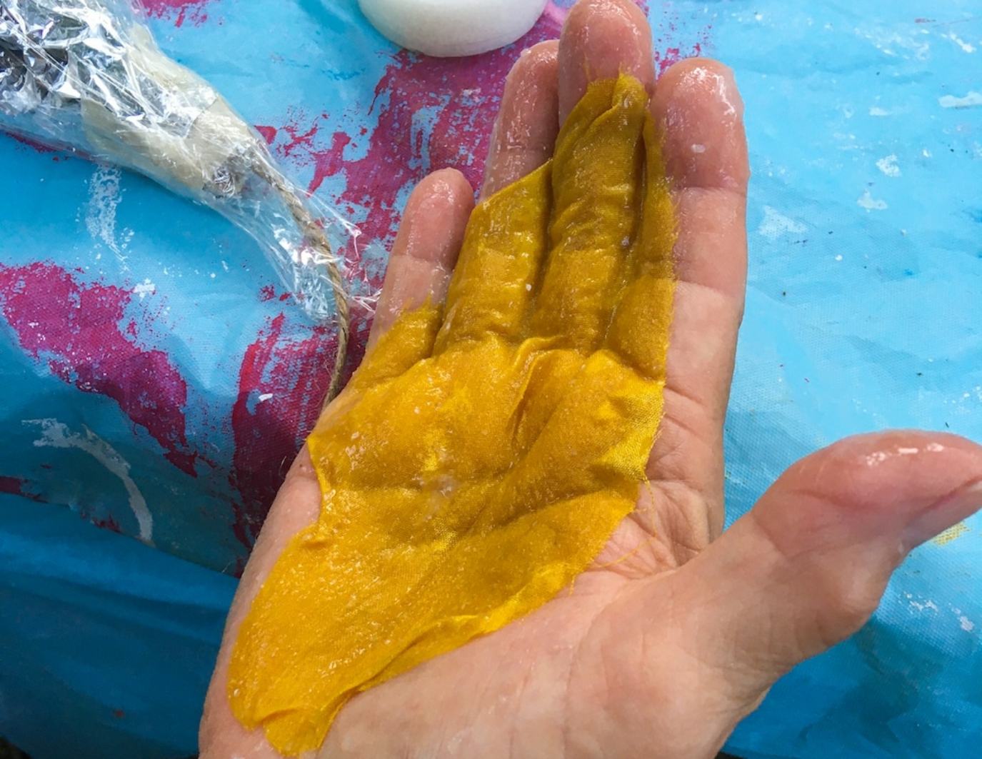 A yellow hand dyed flower is in the right palm of a hand, facing up. The flower is sort of see through, and appears wet, as if just recently dyed. A blue table is the background, with splashes of pink.