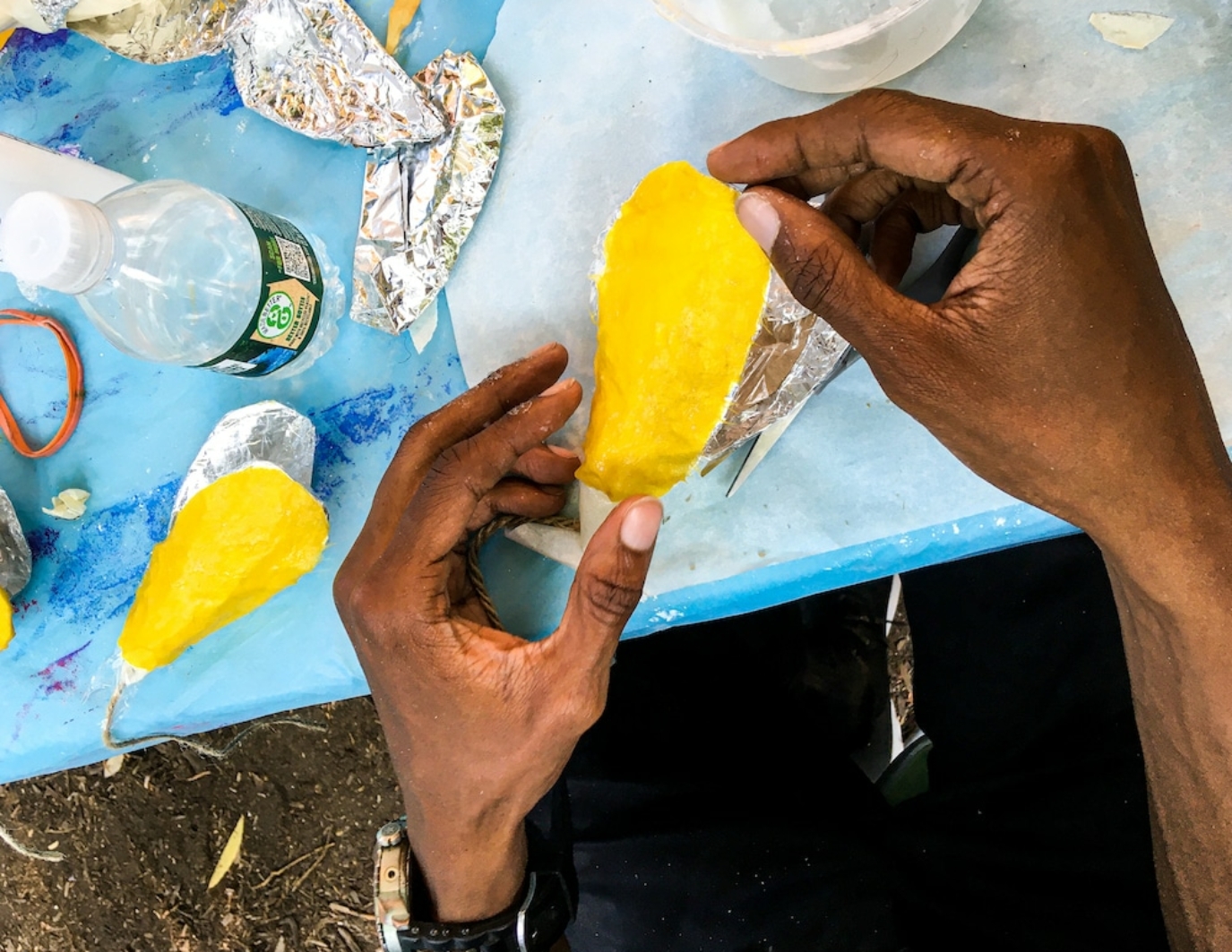 One pair of hands comes up from the bottom of the image and is holding a yellow flower that sort of looks like a mango. The blue table the hands rest on is filled with materials and tinfoil. This is the NYC event.
