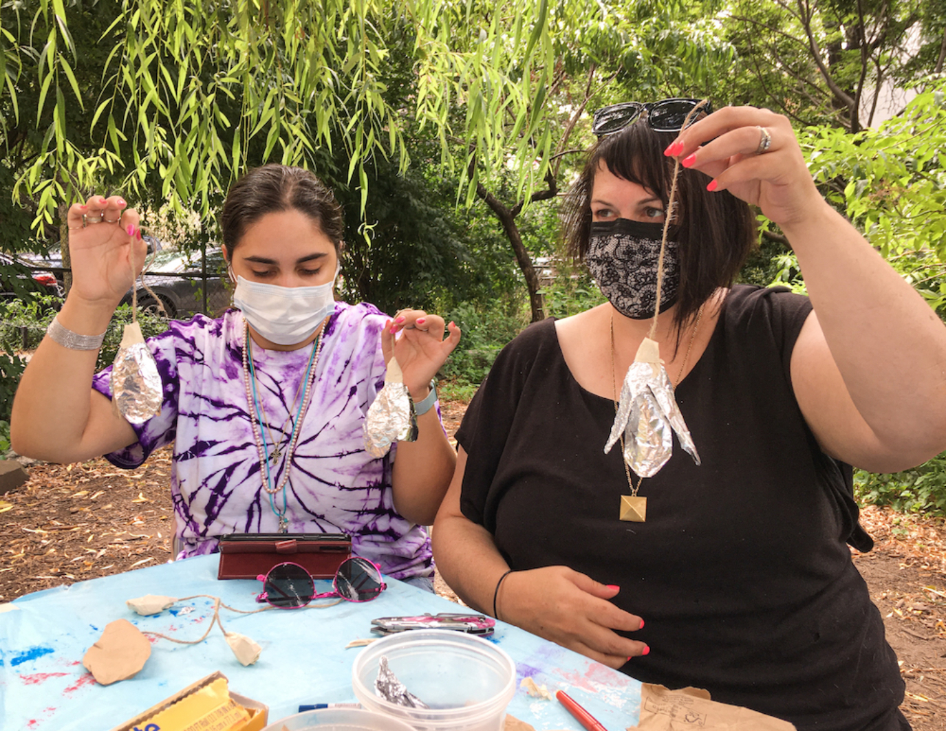 Two volunteers are masked and seated and holding up materials they've made. The left person has a ty dyed shirt of purople and is holding up two ornaments of foil that appear as flowers, while the person on the right is in black shirt and holding one ornamental in their left hand. They are seated around blue work table, are outside, due to COVID safety, and are masked. There are trees surrounding them. This is in NYC.
