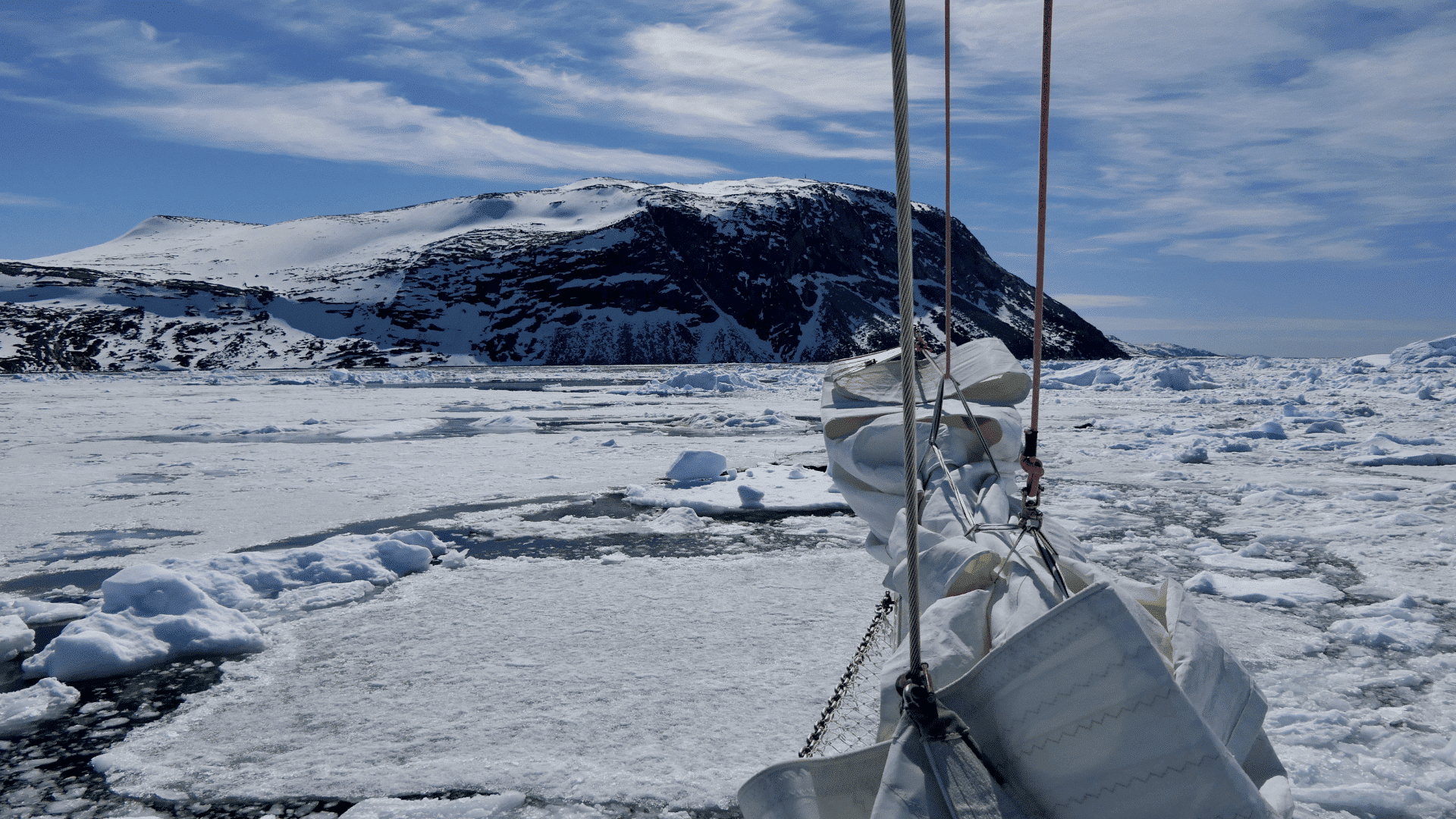 The bow of a boat as it cuts through water covered in snowy ice. Rocky land looms in the distance.